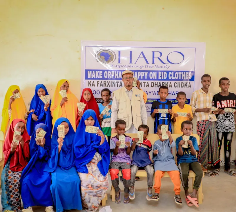 Children in new Eid clothes hold their gifts beside HARO staff under the Make Orphans Happy on Eid banner